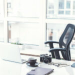 Modern office. Empty workplace with white laptop camera, coffee cup and other items. Big window with cityscape on background