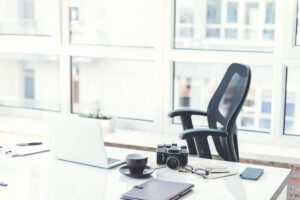 Modern office. Empty workplace with white laptop camera, coffee cup and other items. Big window with cityscape on background