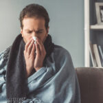 Handsome sick man is wiping nose while sitting on couch covered in plaid