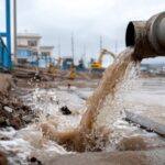 The image captures a water pipe actively discharging muddy water into a flooded urban area, highlighting infrastructure challenges during rainy weather.